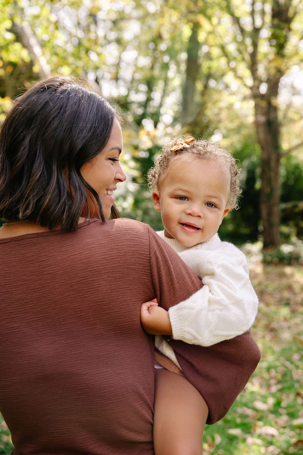 A smiling mother holds her cute toddler girl outdoors in a lush green park setting.