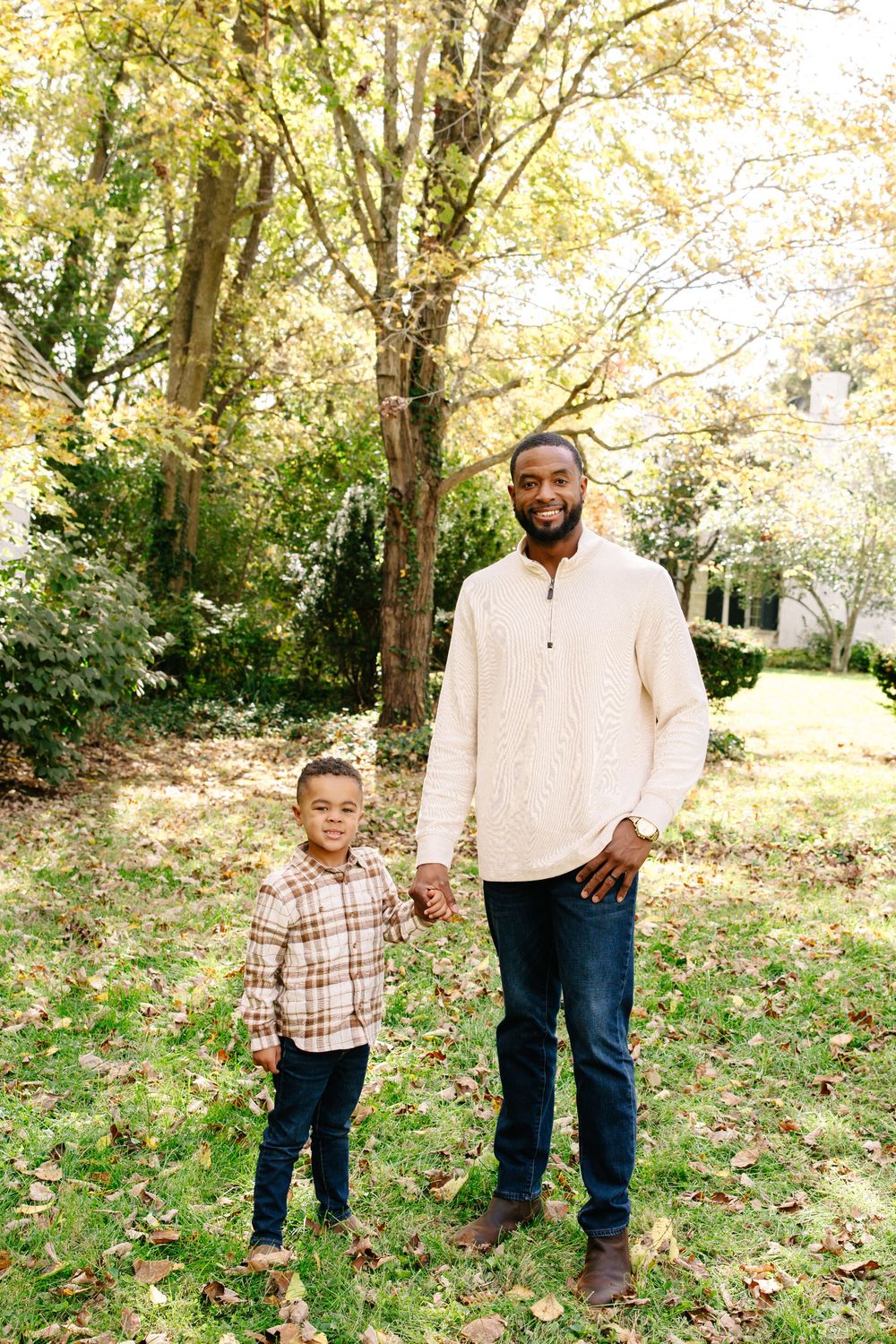 A smiling father and young son hold hands outdoors in a fall park setting, both wearing casual earth-tone outfits.