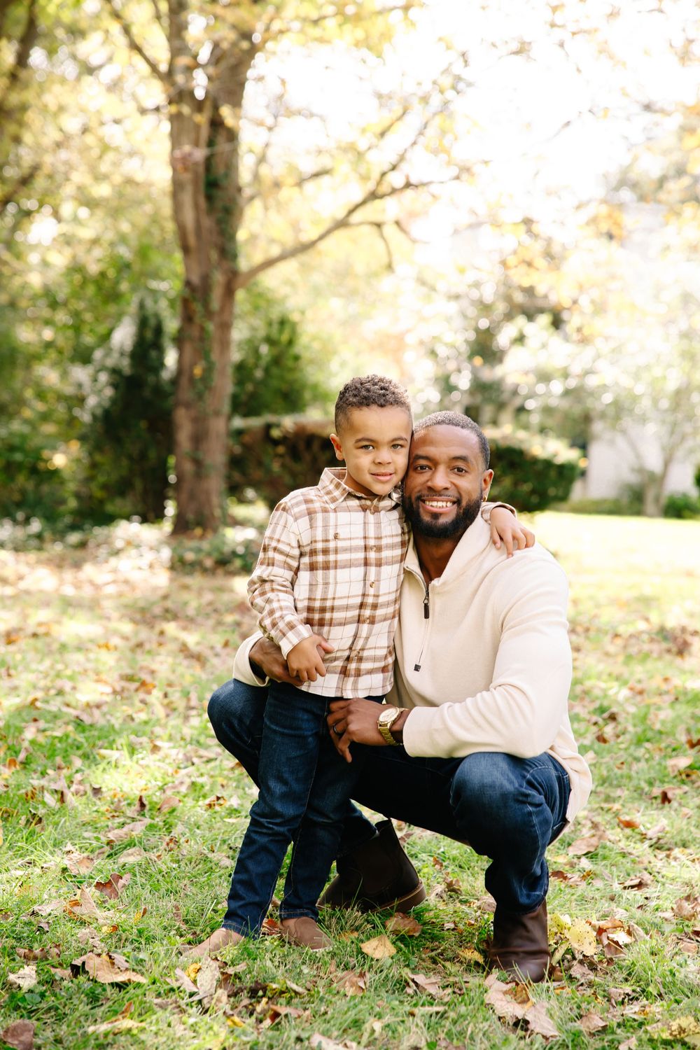 A smiling man crouches beside a young boy in a sunny park, both wearing casual fall outfits among autumn leaves.