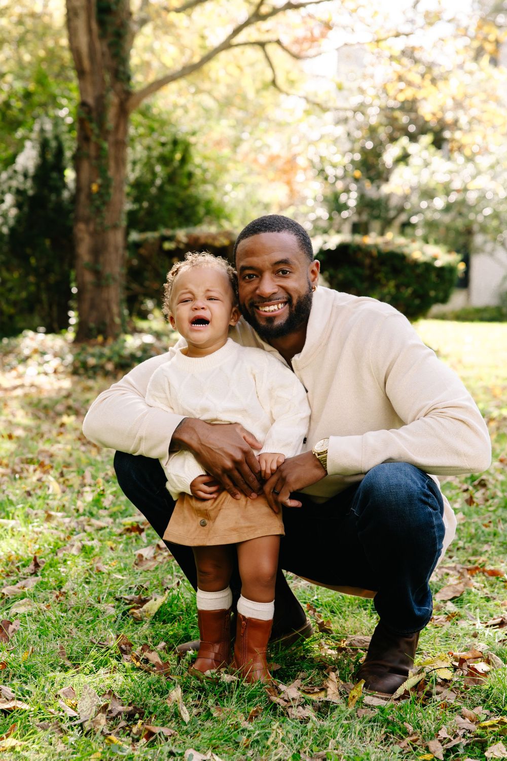A smiling man crouches beside a laughing toddler girl in a lush green park during autumn.