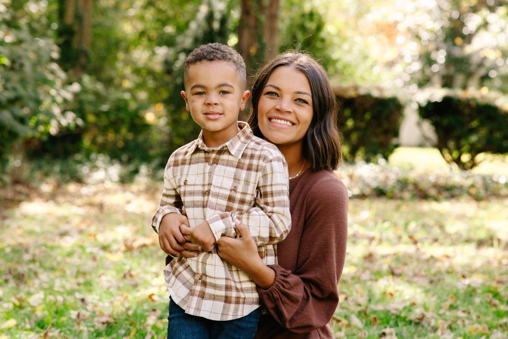 A woman smiling and holding a young boy outdoors in a park setting with green trees in the background.
