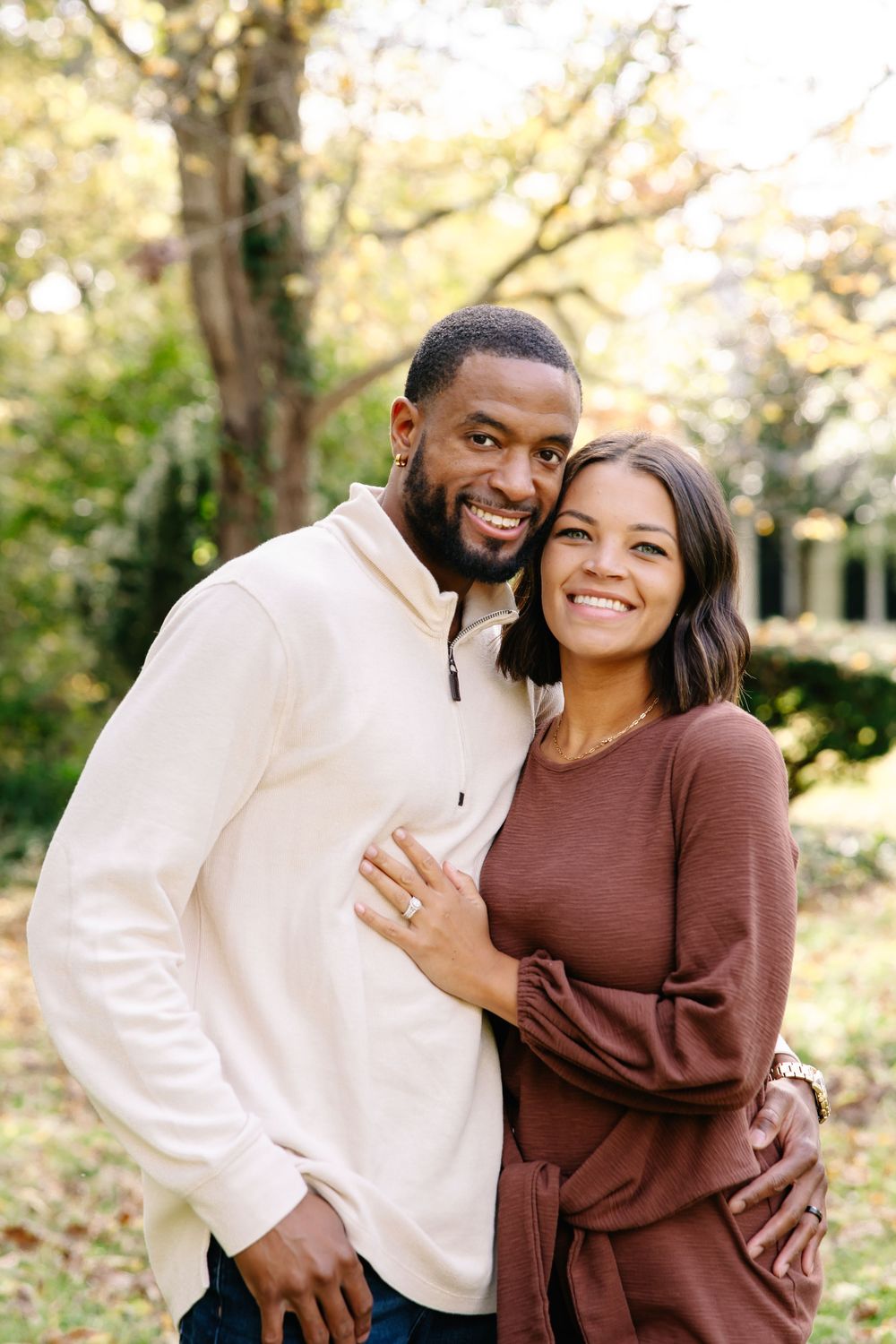 Smiling couple embracing outdoors in autumn, man in cream pullover and woman in burgundy top, trees in background.