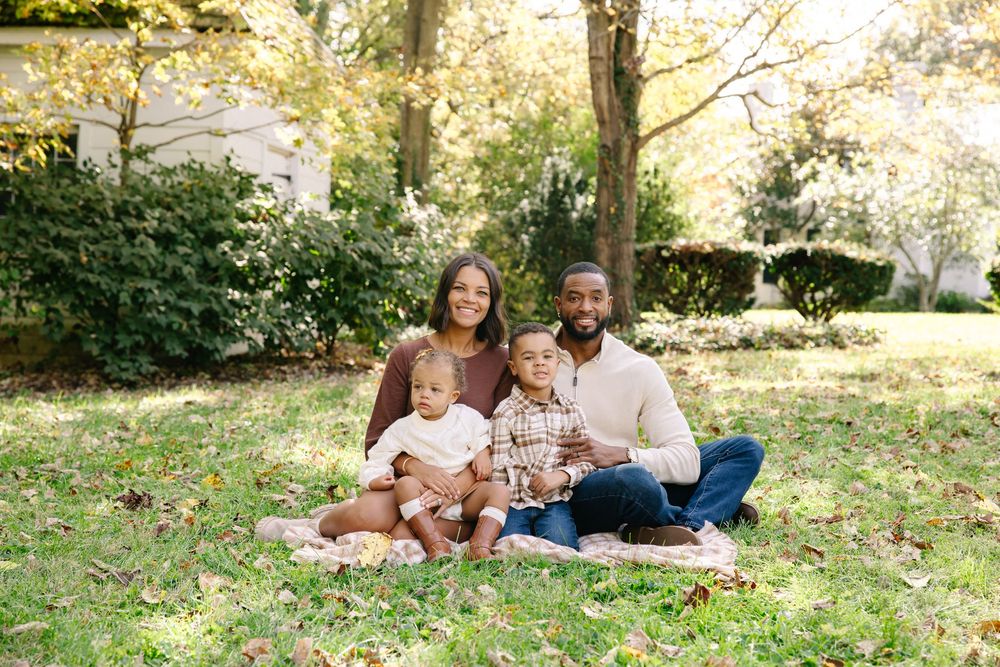 Happy family of four sitting on grass in a sunny park during autumn, with two young children and smiling parents.