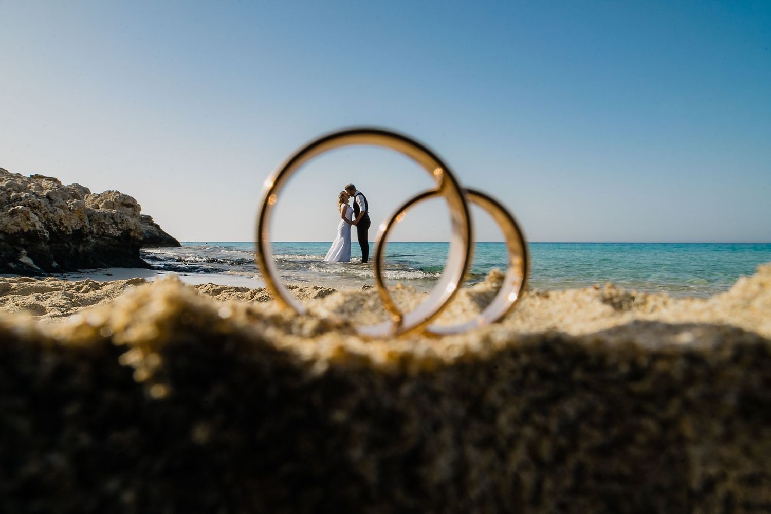 Two gold wedding rings on a sandy beach with a couple kissing by the ocean in the background.