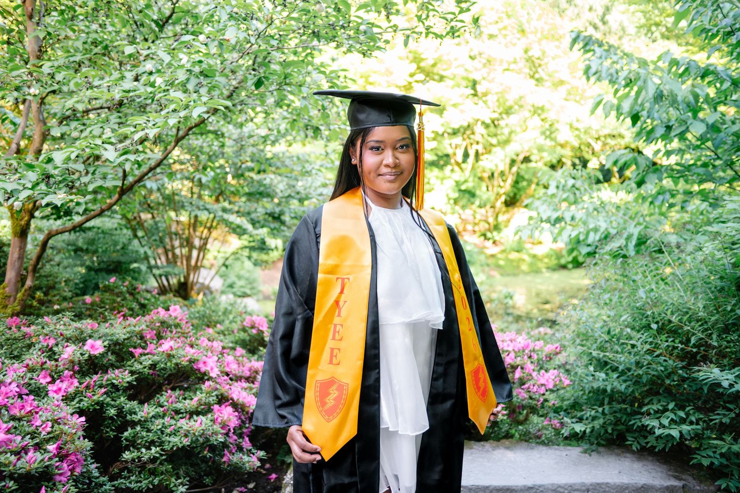 Graduate wearing a cap and gold stole among the flowers and greenery of the Yao Garden in Bellevue