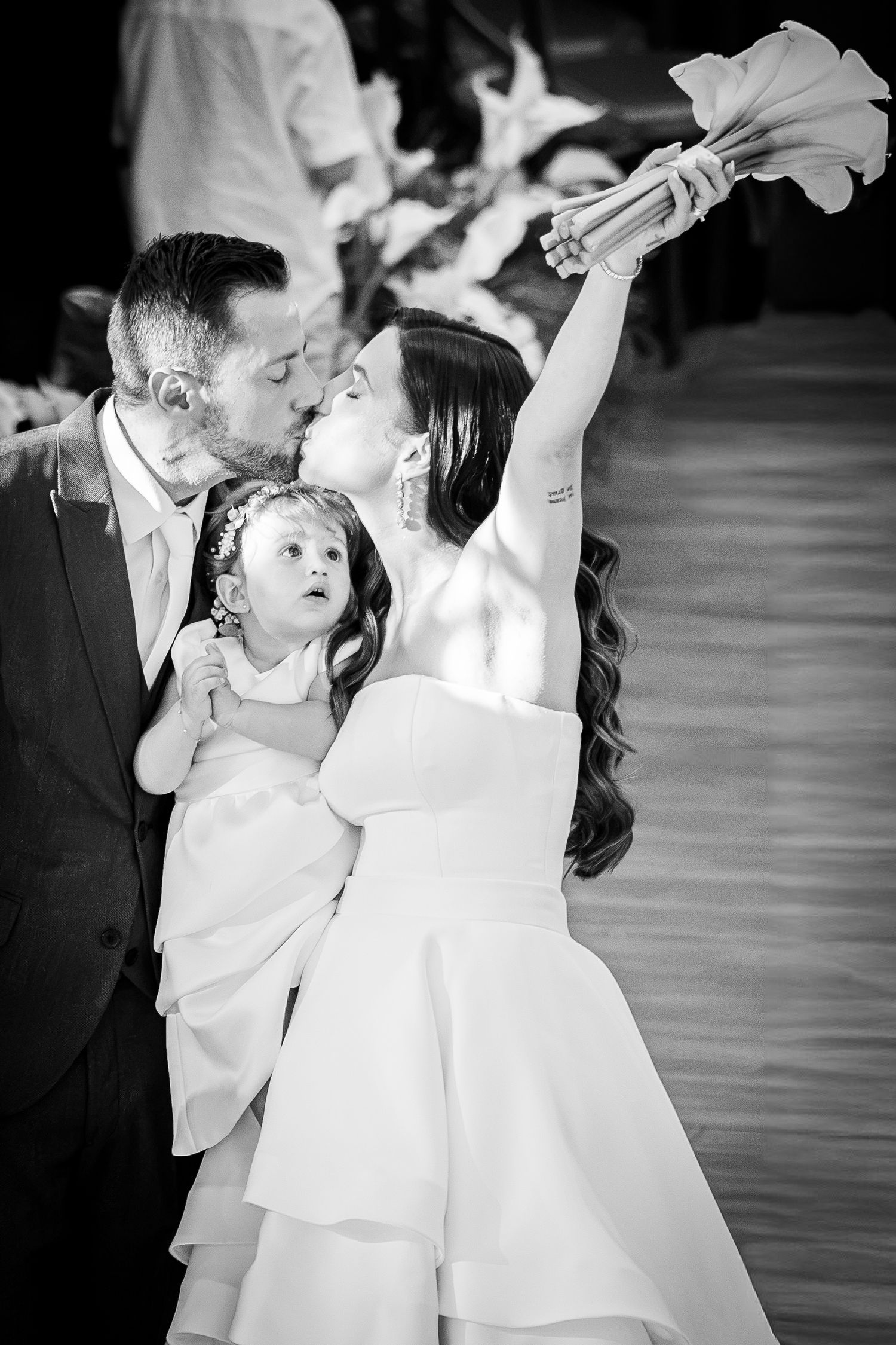 Black and white wedding photo of bride and groom kissing while holding a young flower girl, bride raising bouquet.
