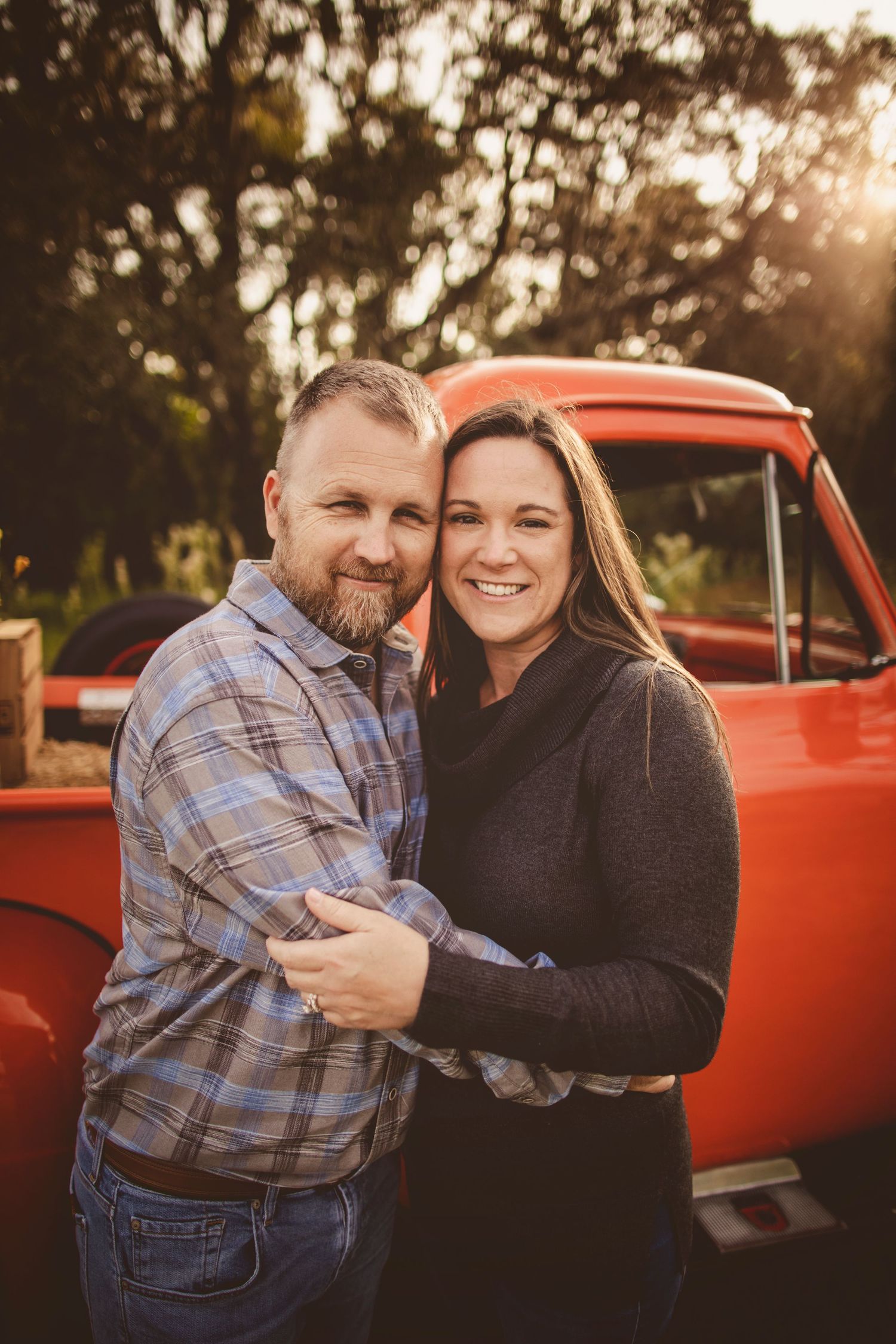 Smiling couple embracing outdoors near a vintage red truck with golden sunset light filtering through trees.