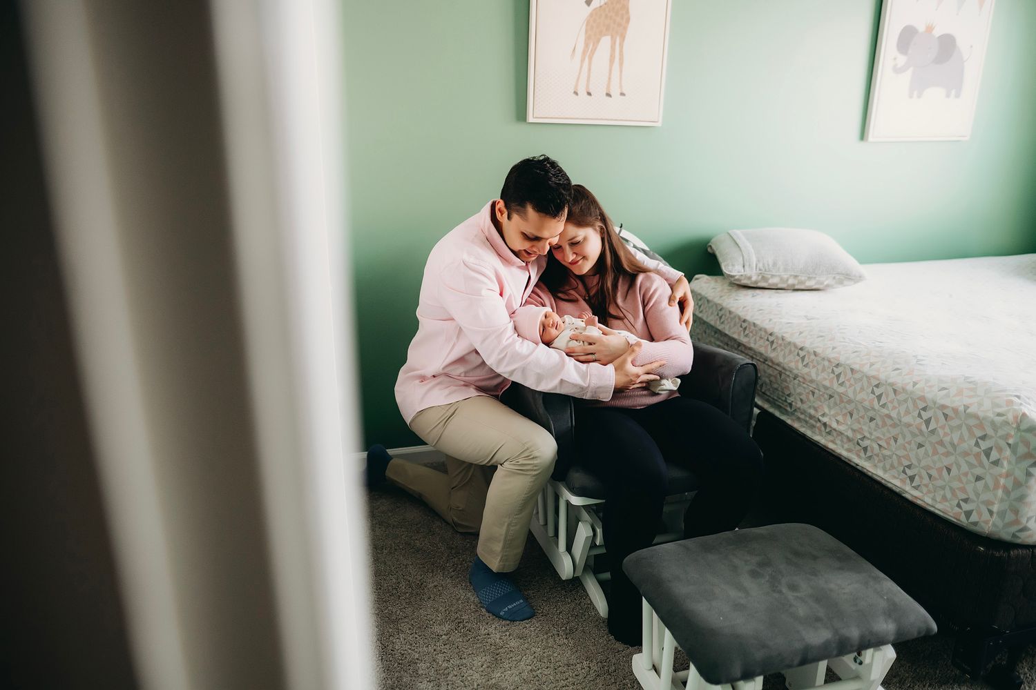 A couple sits together on a bed, lovingly cradling their newborn baby in a softly decorated nursery room.