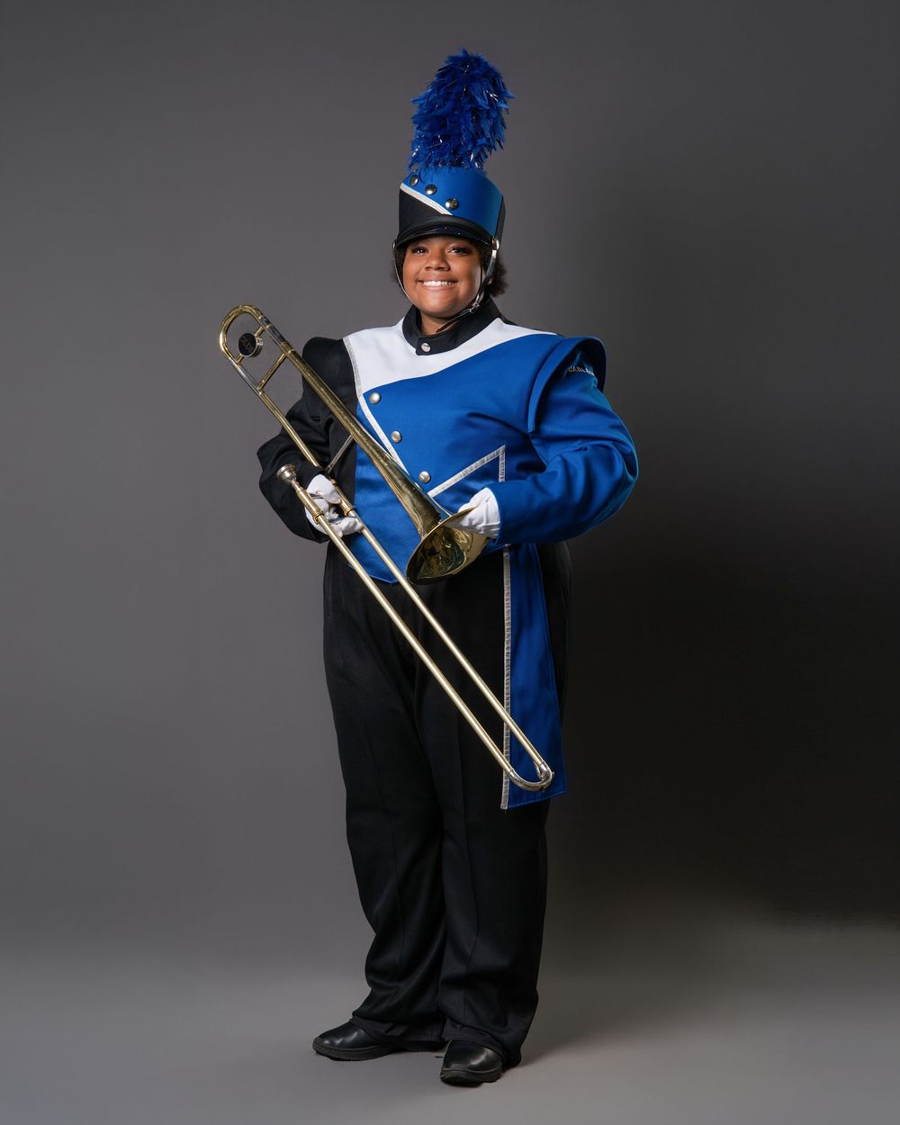 A smiling marching band member in a blue uniform holds a trombone, wearing a blue feathered shako hat.