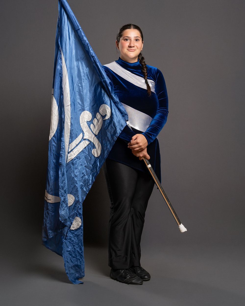 Young woman in blue velvet uniform holds a large blue and white marching band color guard flag against a gray backdrop.