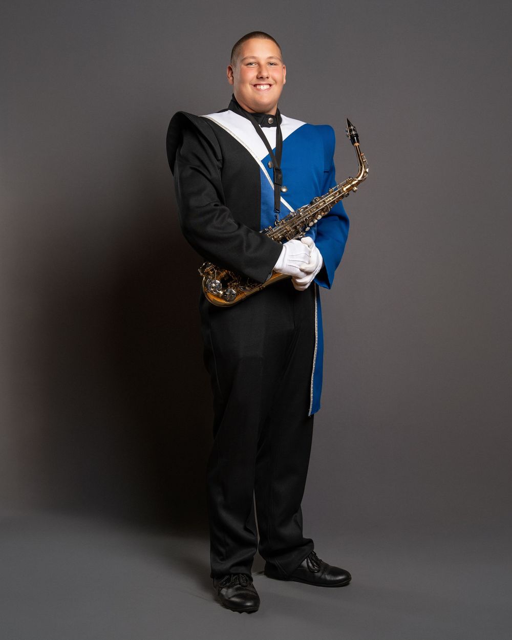 Smiling man in blue and black marching band uniform holding a saxophone, wearing white gloves, posed against gray backdrop.