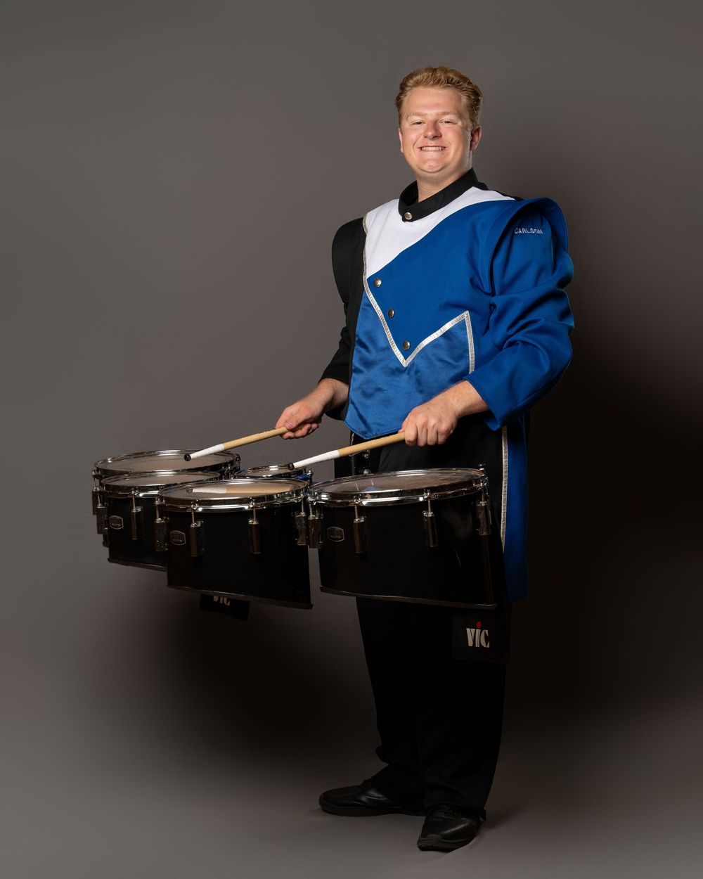 Marching band drummer in blue and white uniform playing tenor drums with drumsticks on gray background.