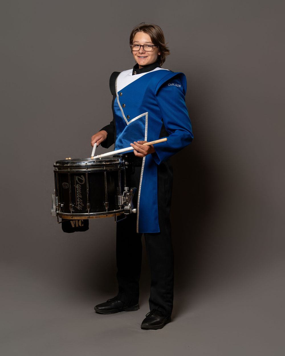 A young marching band drummer in a blue and white uniform holds drumsticks and a snare drum, posing for a portrait.