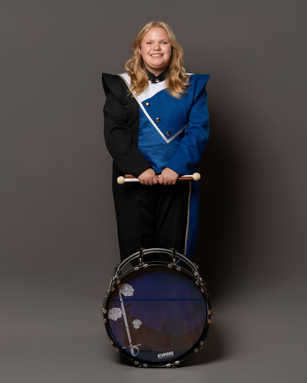 Student in blue and black marching band uniform holds drumsticks over a bass drum in a studio portrait.