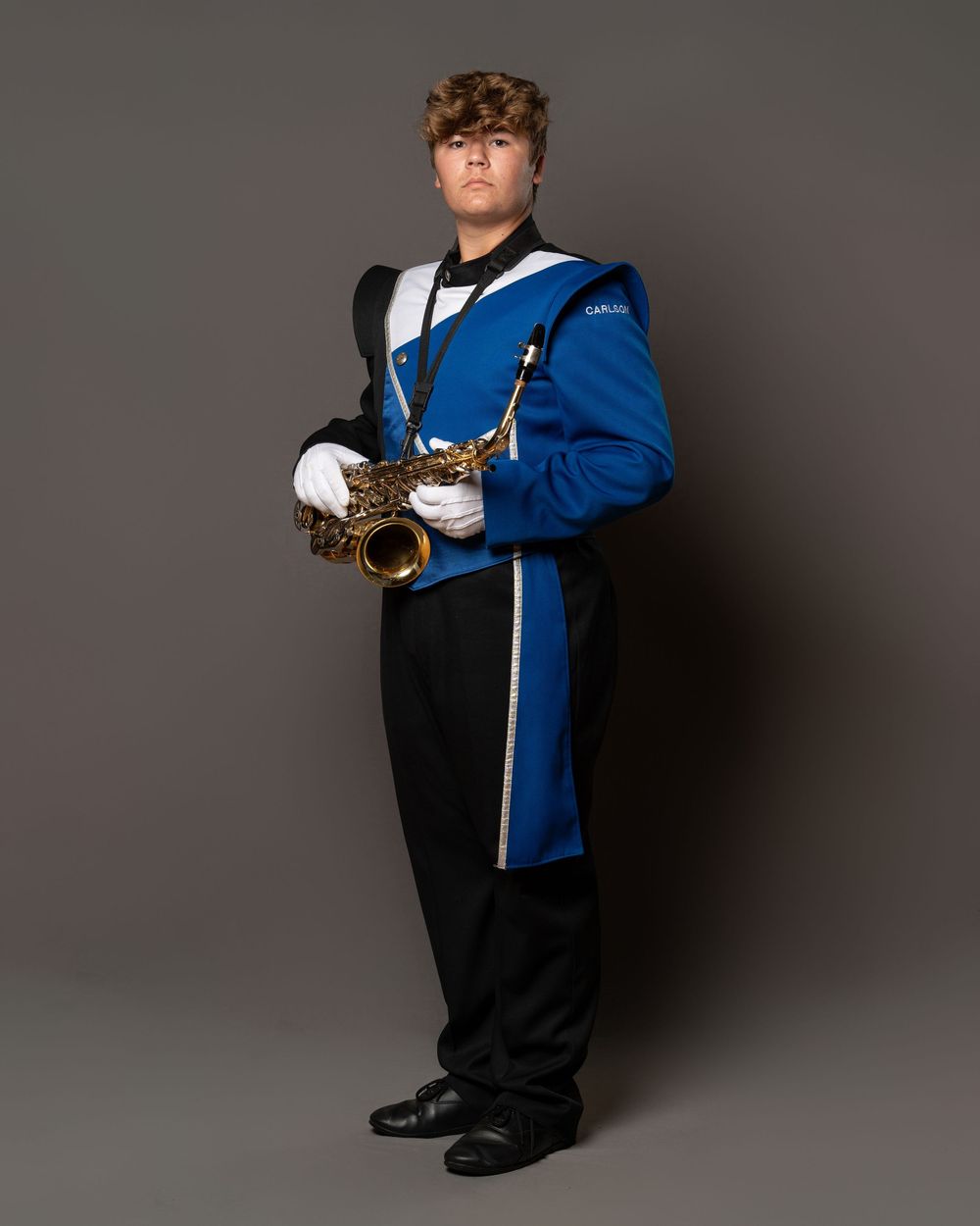 Student in blue marching band uniform holds saxophone, wearing white gloves and black pants for a formal portrait.