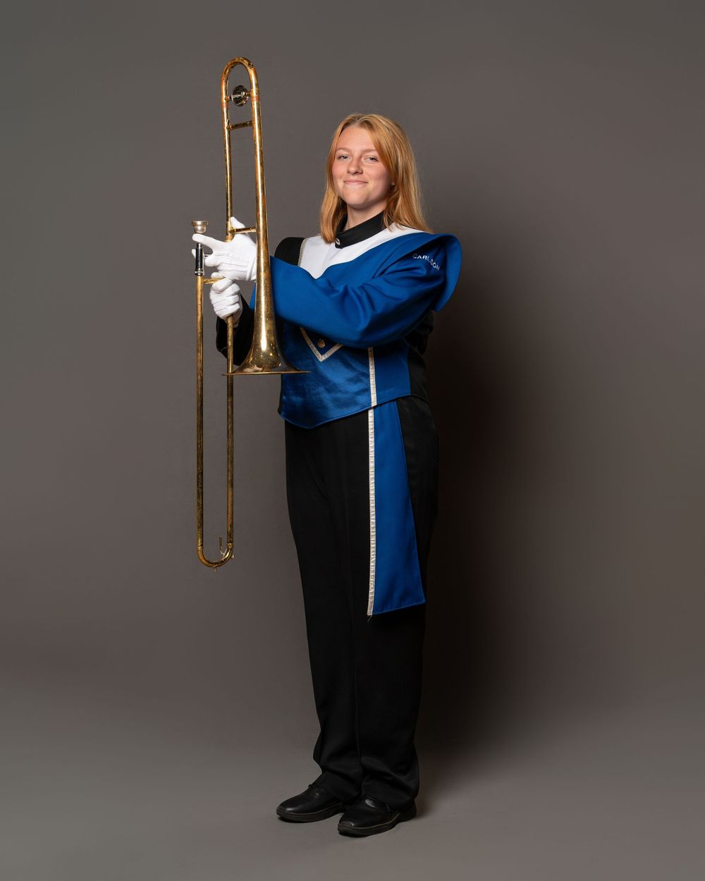 Marching band member in blue uniform holding a trombone, wearing white gloves, posing for a portrait.