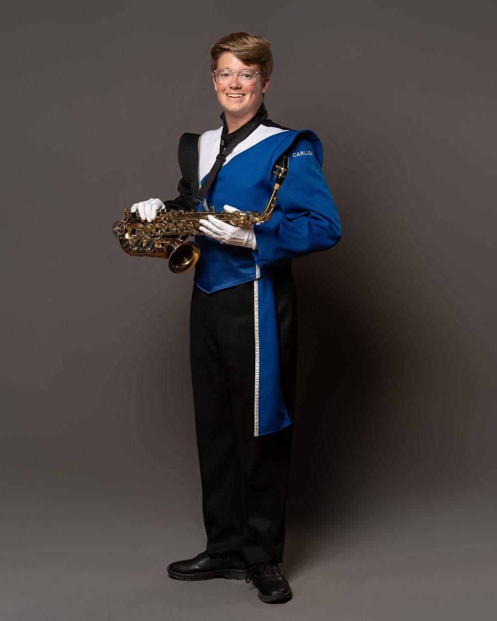 Marching band member in blue uniform holding alto saxophone, wearing white gloves and black pants, posing for portrait.