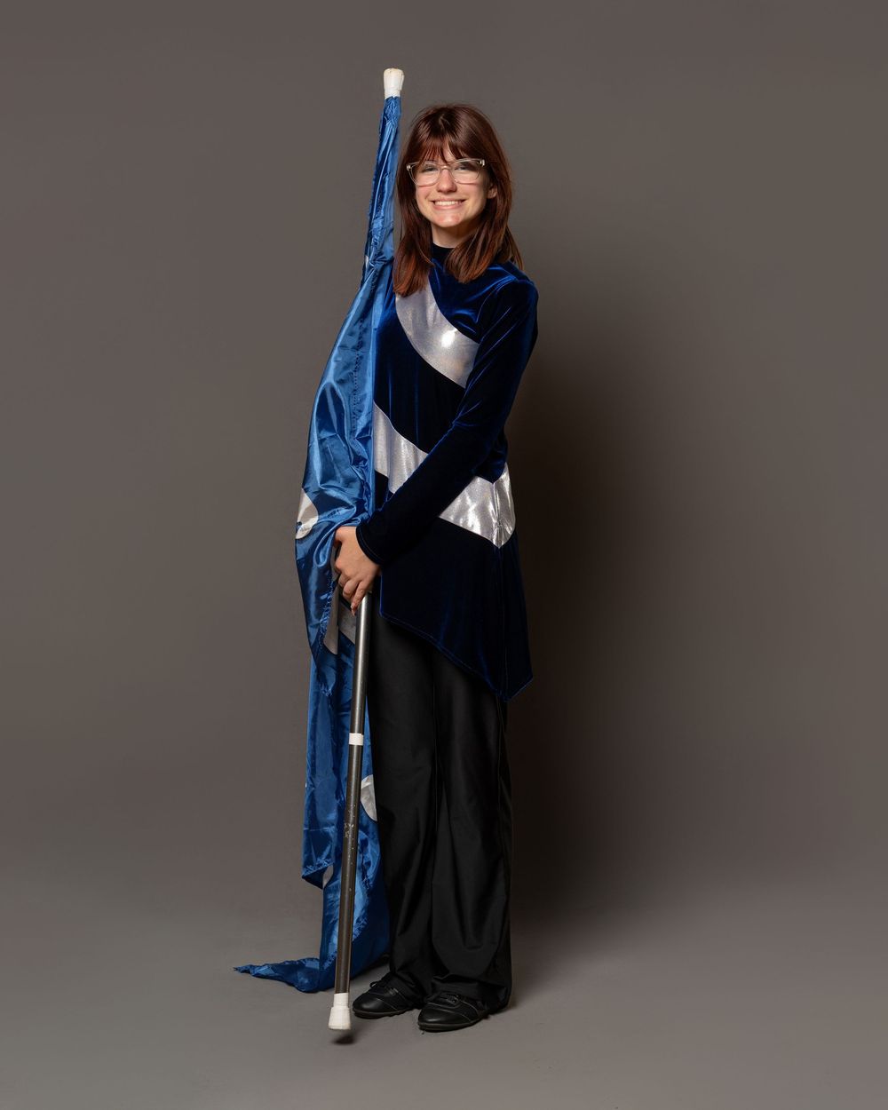 Young woman in blue and silver marching band uniform holding a color guard flag pole, posing on gray background.