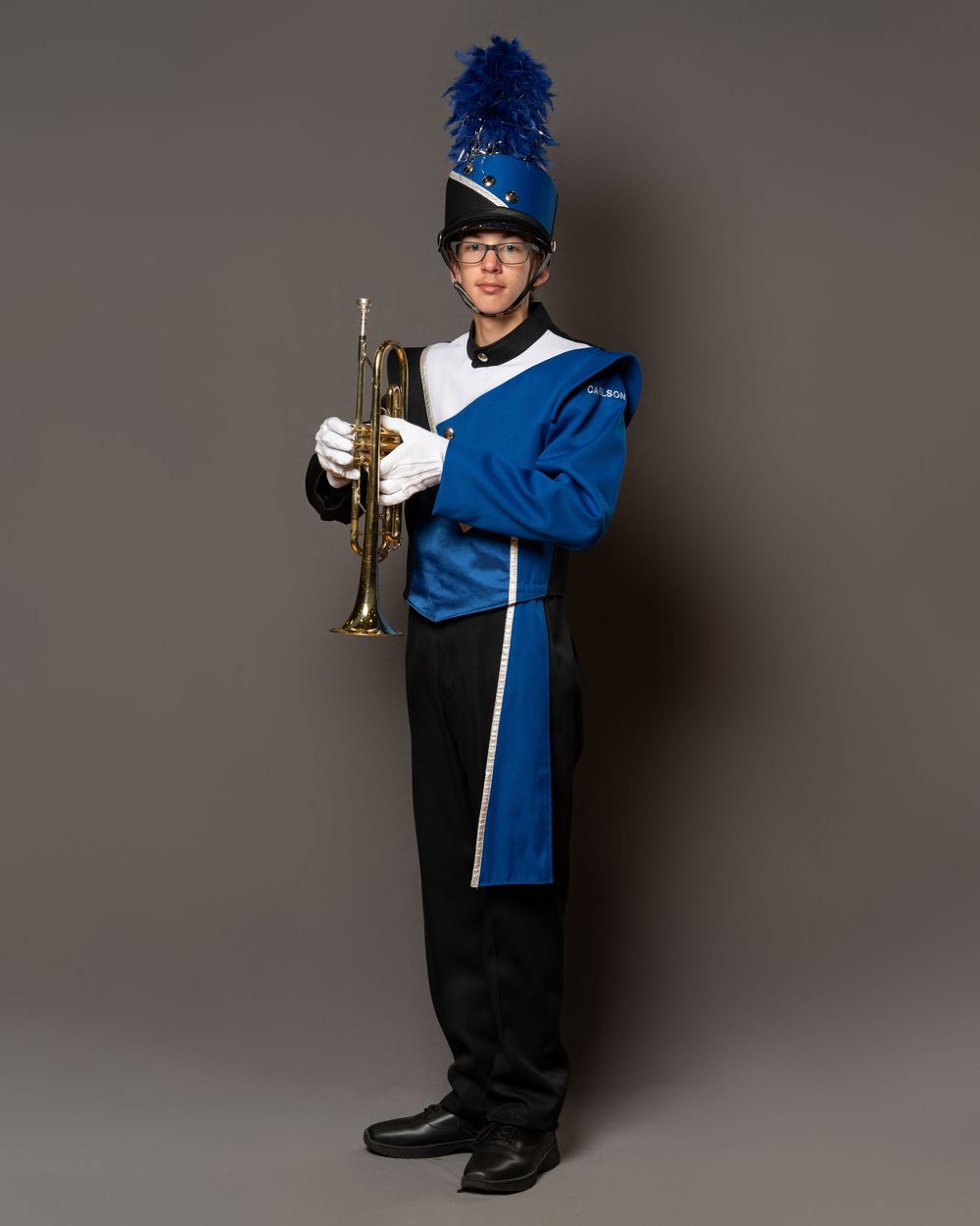 Marching band member in blue uniform holding a trumpet, wearing a feathered shako hat and white gloves.