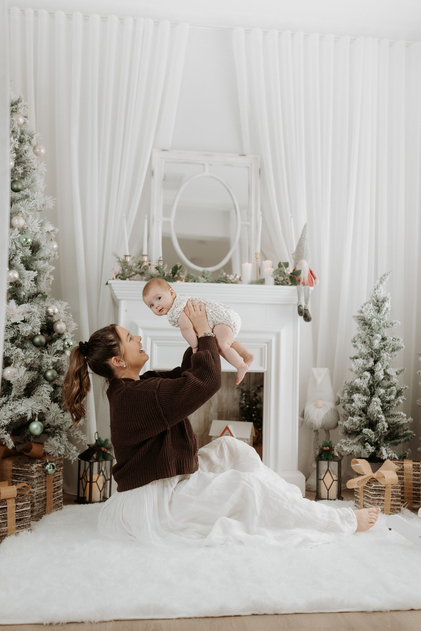A mother joyfully lifts her baby near a white fireplace with snowy Christmas trees and gifts in a festive holiday setting.