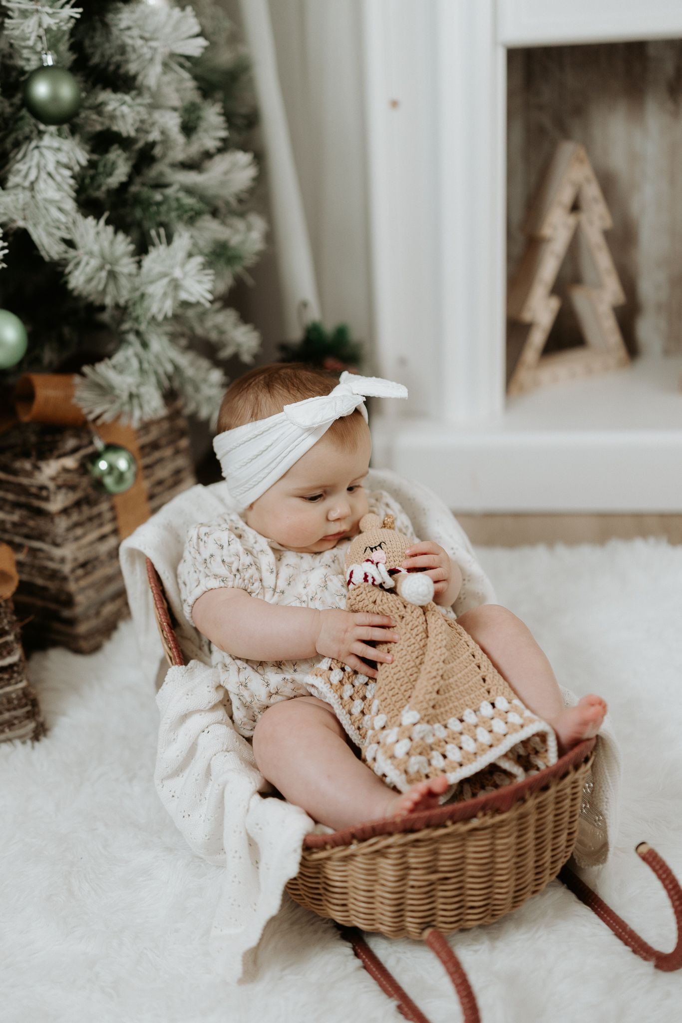 Baby girl in white headband sits in wicker basket holding crochet toy near Christmas tree on fluffy white rug.