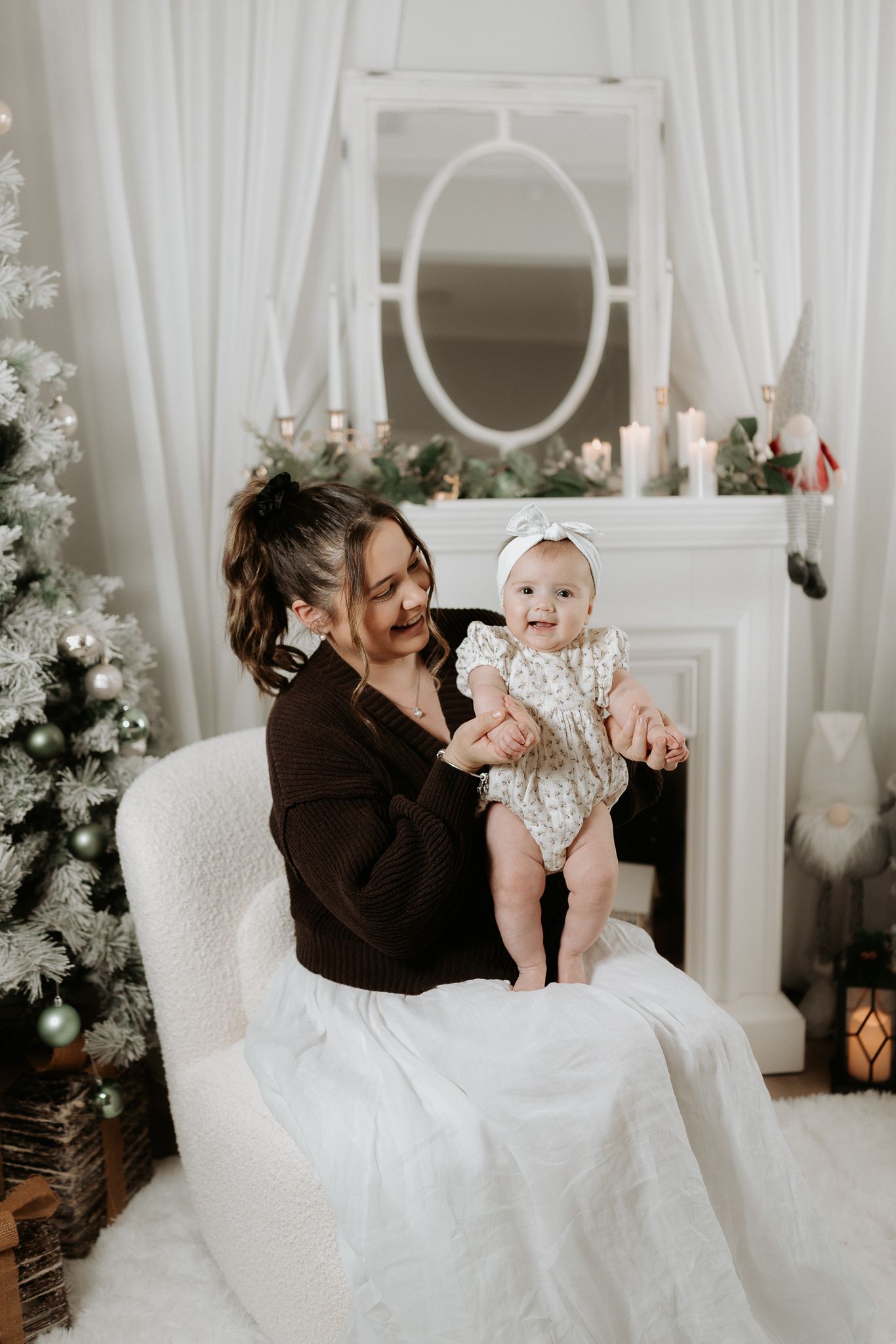 A mother holds her baby girl in a white chair beside a Christmas tree and decorated fireplace mantel.