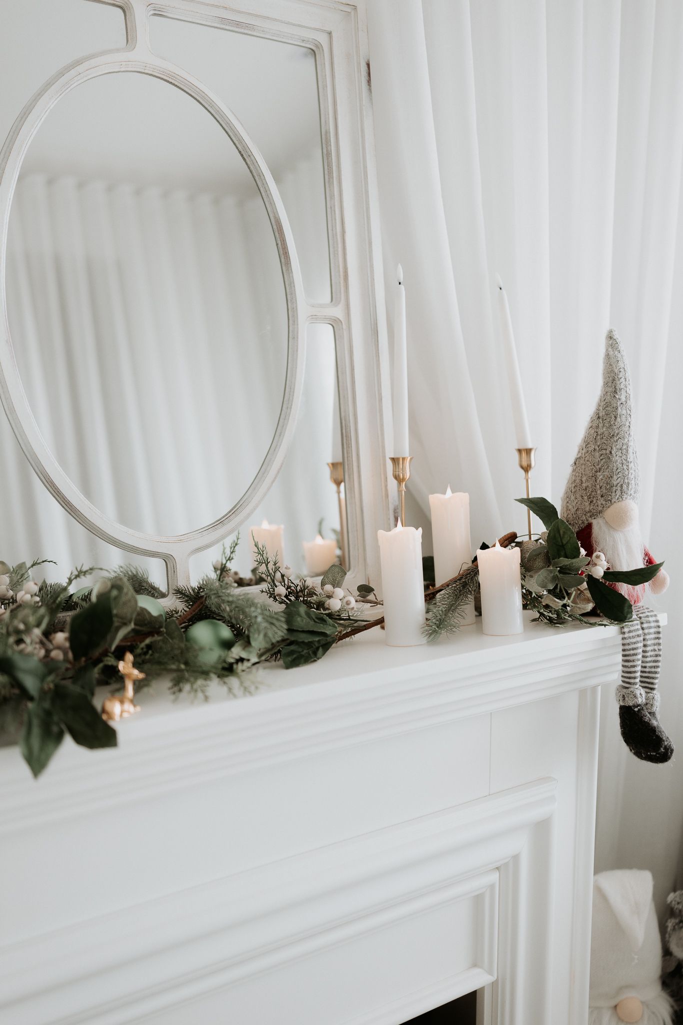 White fireplace mantel with Christmas garland, white candles, and a gnome figurine beneath an oval mirror.