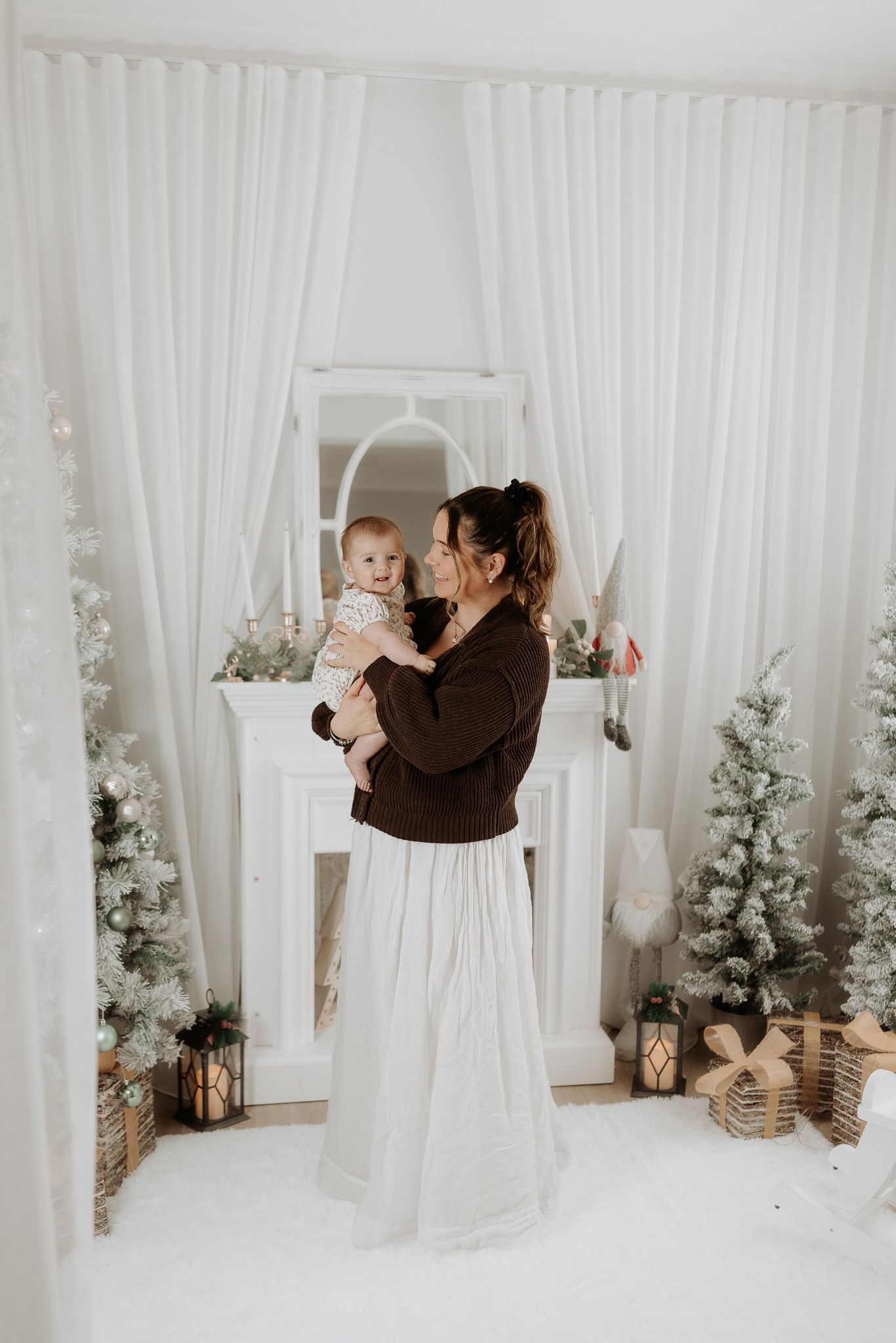 A mother in a brown sweater and white skirt holds her baby near a white fireplace with snowy Christmas trees and gifts.