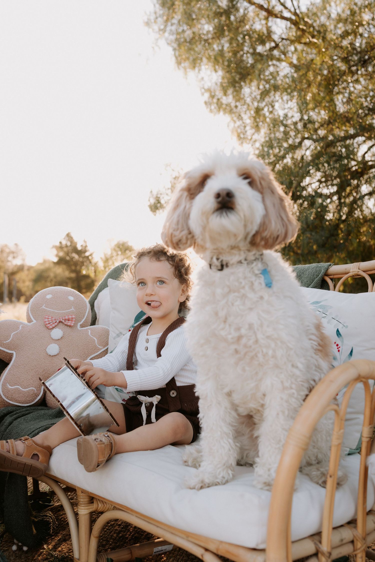 A young child sits on a rattan bench with a fluffy white dog and a gingerbread man pillow outdoors.