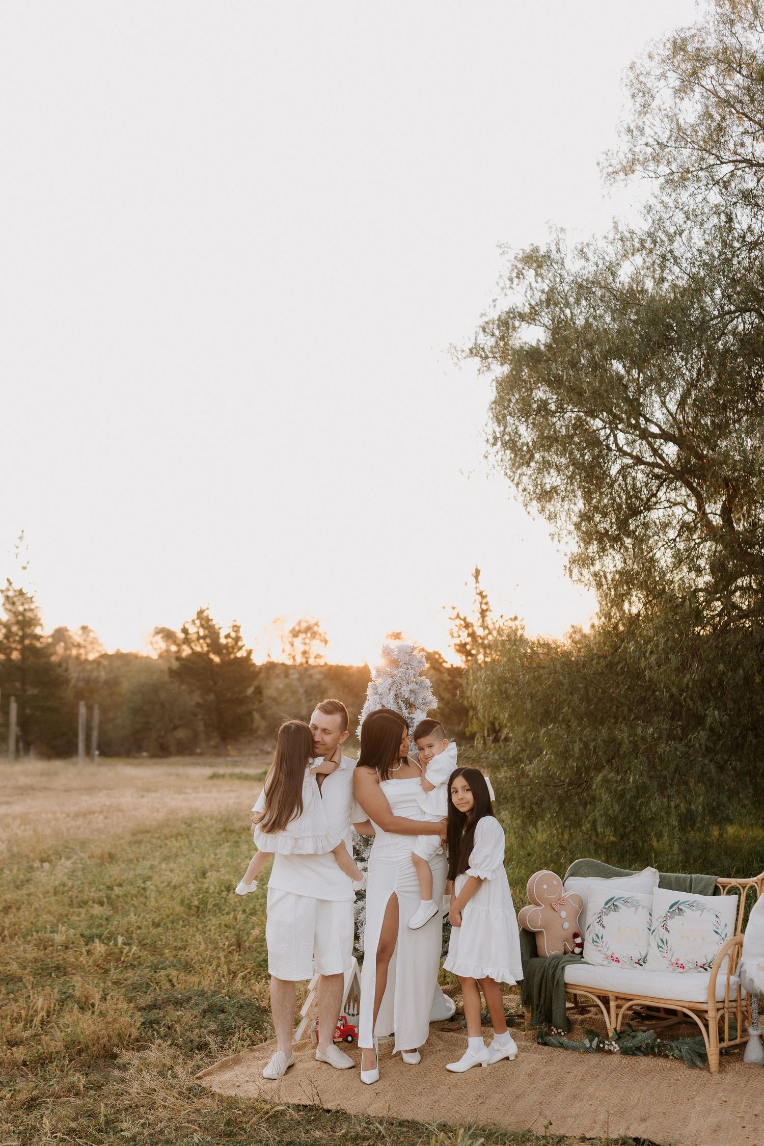 Family in white outfits embracing outdoors at golden hour near a rattan sofa in a sunlit field.