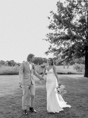 Black and white photo of a bride and groom holding hands on a grassy lawn with a large tree backdrop.
