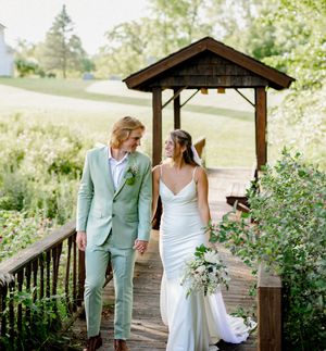 A bride and groom walk hand-in-hand on a wooden deck surrounded by lush greenery on their wedding day.