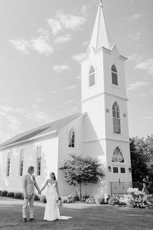 Black and white photo of bride and groom holding hands in front of a white chapel with a steeple.
