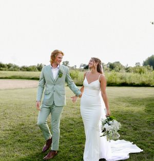 A bride in a white gown and groom in a sage green suit hold hands while walking on a lush green lawn.