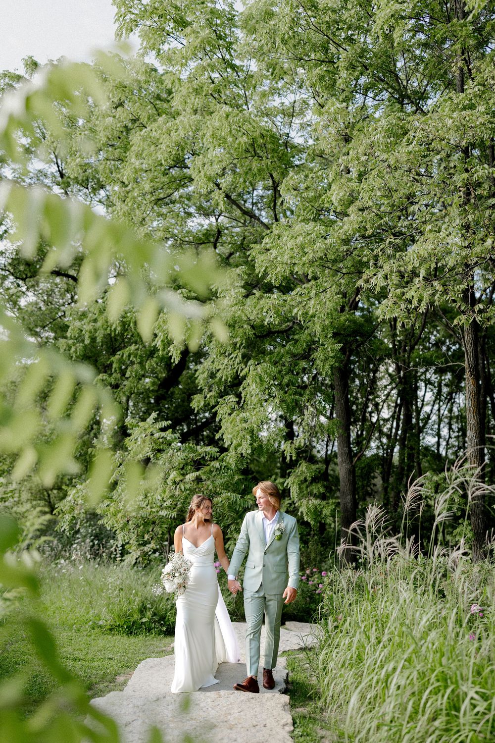 Bride and groom walk hand-in-hand on a garden path surrounded by lush green trees and ornamental grasses.