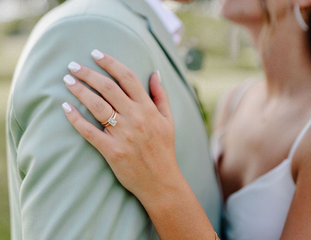 Close-up of a bride's hand with a gold engagement ring resting on a groom's sage green suit jacket.