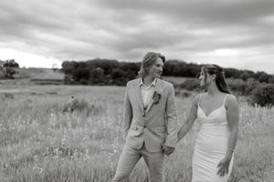 Black and white photo of a couple holding hands in a field on their wedding day, smiling at each other under cloudy skies.