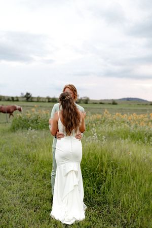 A bride in a white gown embraces her partner in a lush green field with a horse grazing nearby.