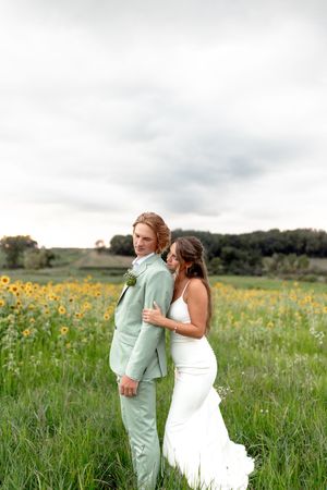Bride in white gown embraces groom in mint suit standing in sunflower field under cloudy sky.