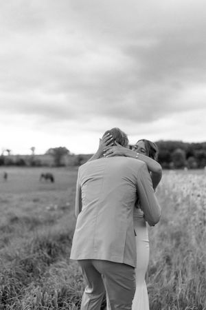 Black and white photo of a couple embracing in a field on their wedding day, with a horse grazing in the background.