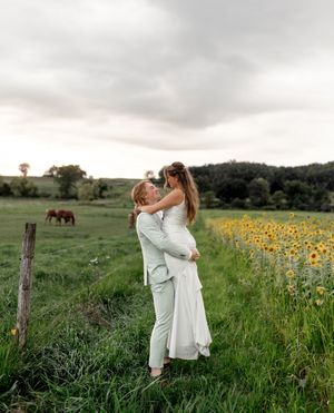 Couple in white attire embracing in a sunflower field with horses grazing in the background.