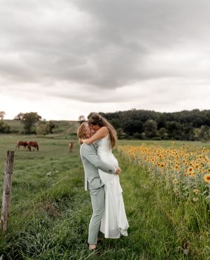 Couple embracing in wedding attire beside a sunflower field with horses grazing in a lush green countryside.