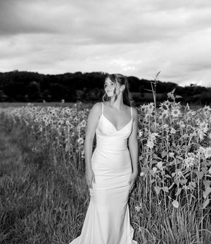 Black and white photo of a bride in a spaghetti strap gown standing in a sunflower field under cloudy skies.