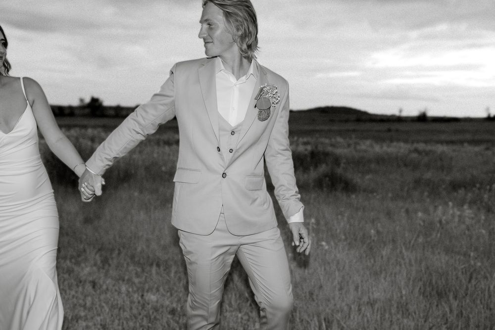 Black and white photo of a couple holding hands in a field, groom in a light suit with floral boutonniere.