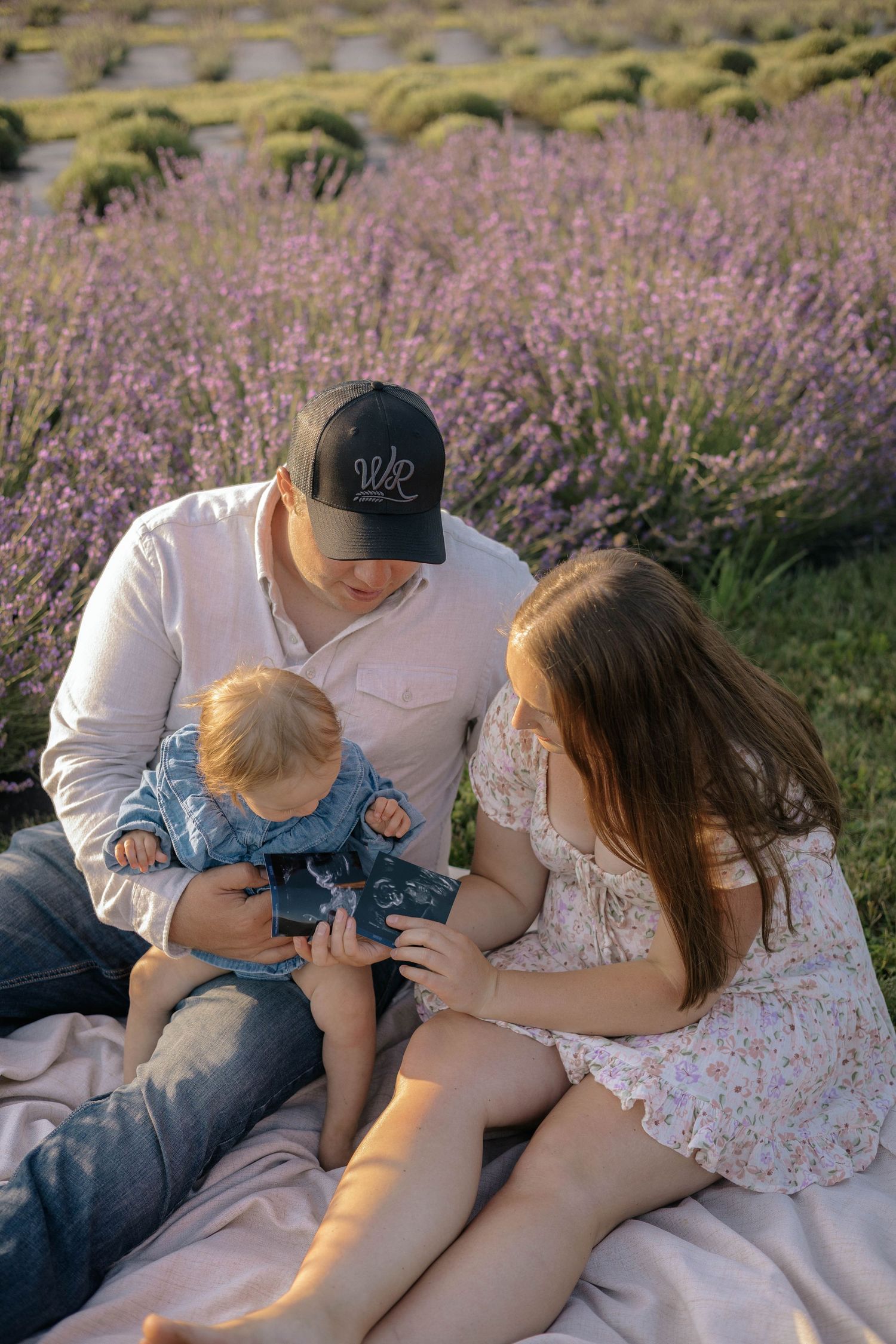 A young family sits in a lavender field, parents lovingly attending to their baby dressed in denim.