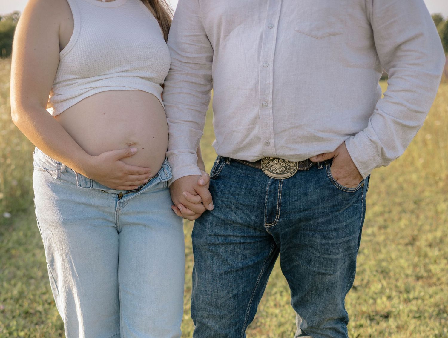 Pregnant couple holding hands outdoors, woman in white top and jeans showing baby bump, man in white shirt and denim.