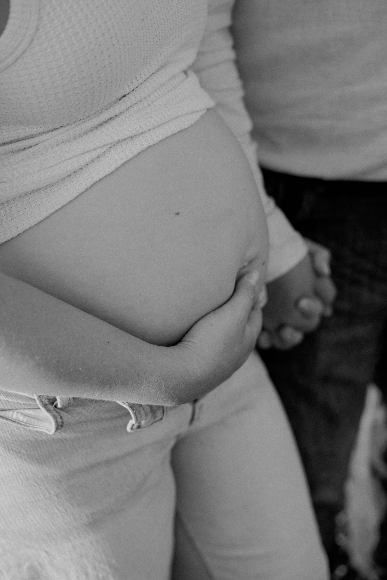 Black and white photo of a pregnant woman cradling her baby bump while holding hands with her partner.
