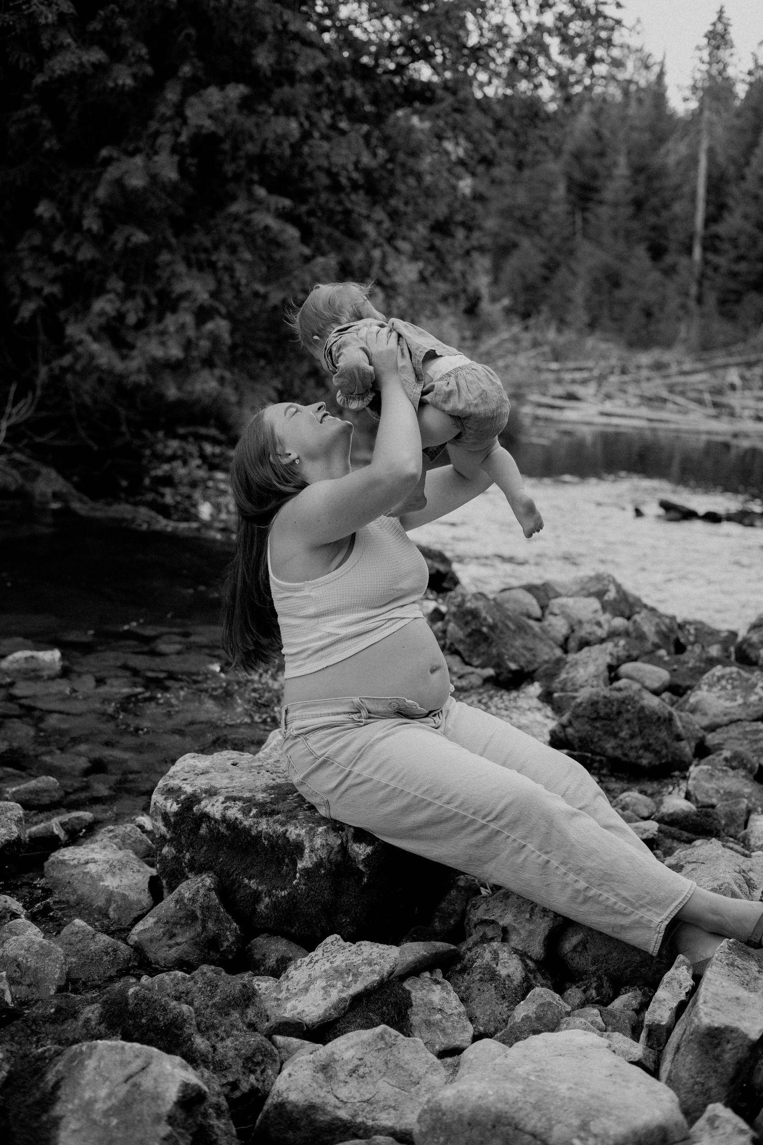 Black and white photo of a mother lifting her baby joyfully while sitting on rocks beside a forest river.