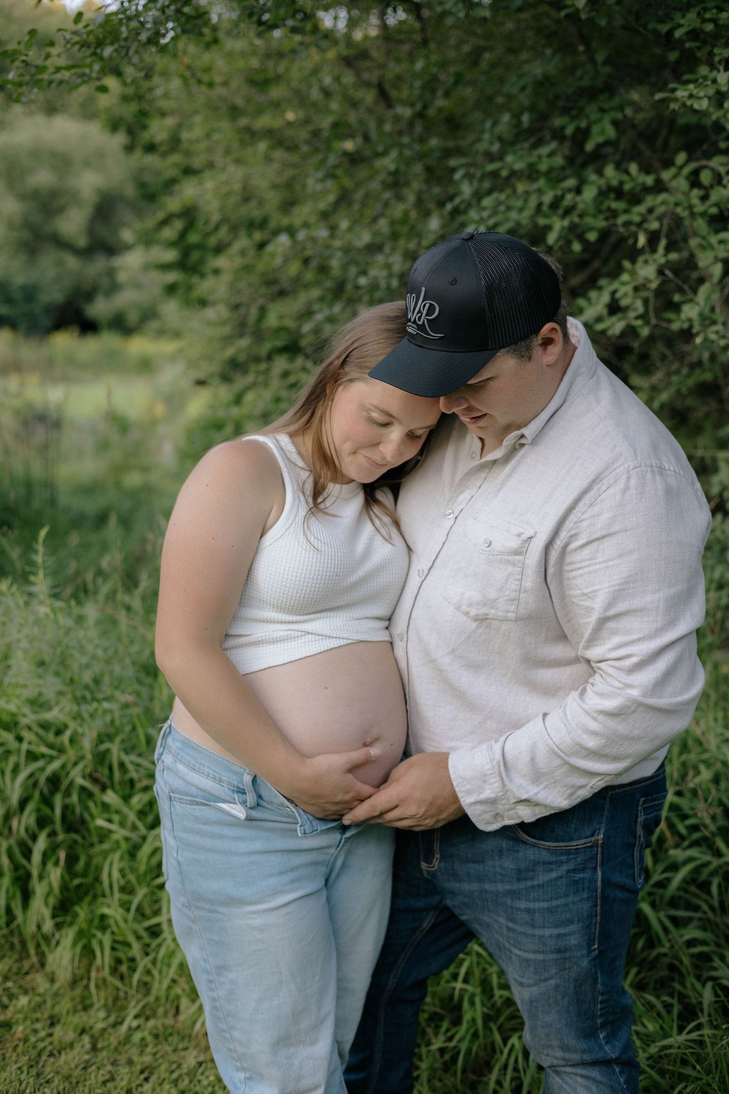 Couple tenderly cradles pregnant belly outdoors, man in cap leans forehead to woman in white crop top and jeans.
