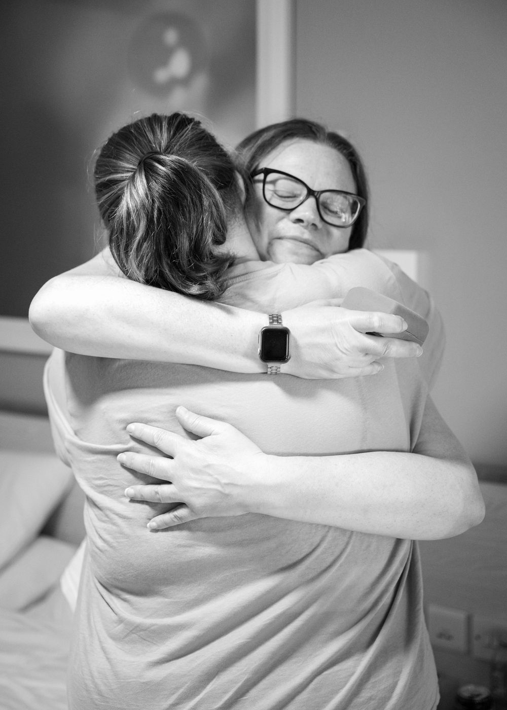Two women share a warm emotional hug in black and white photo, one wearing glasses and an Apple Watch.